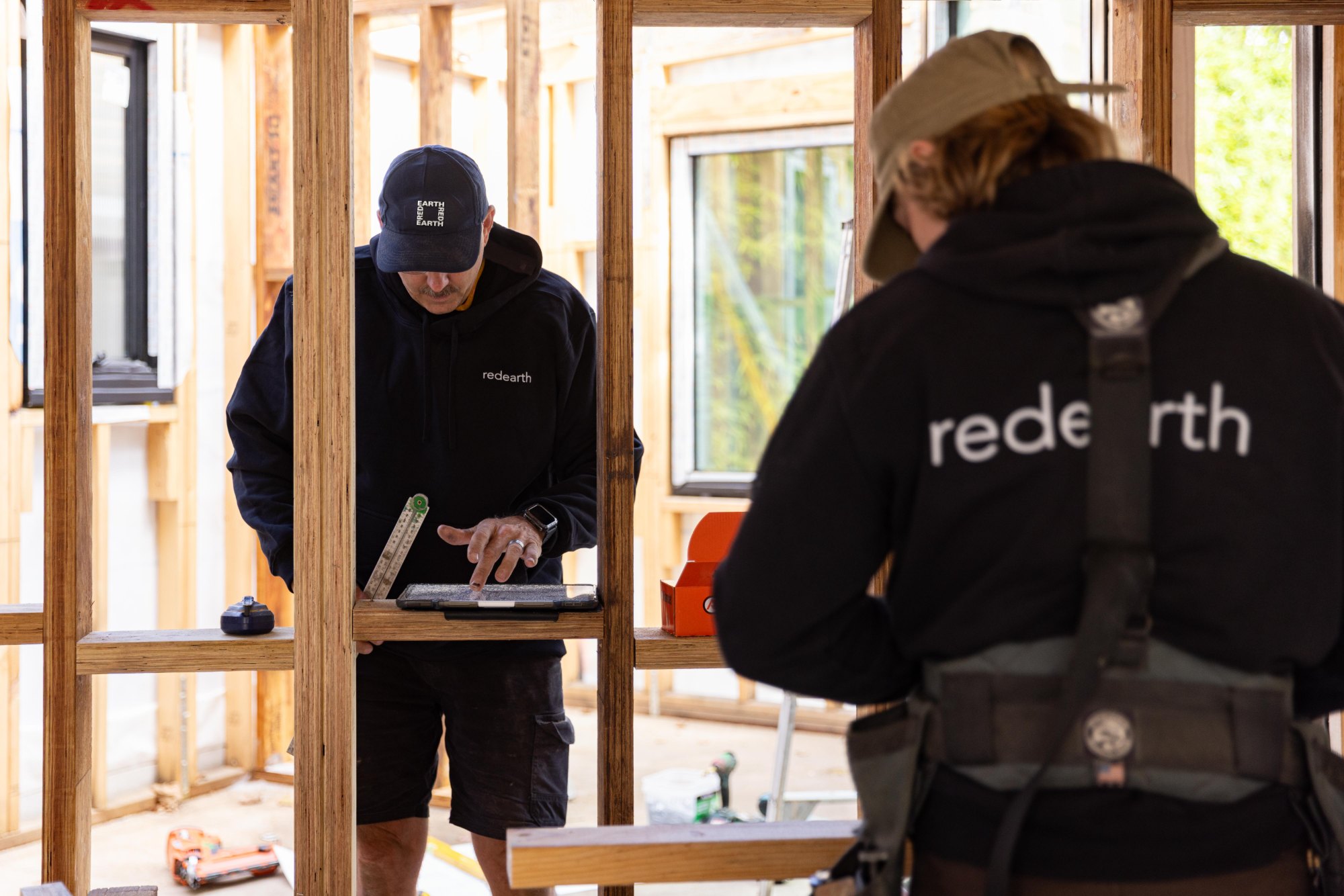Red Earth director checking plans on a tablet at the wall frame, a crew member in a Red Earth hoodie in the foreground